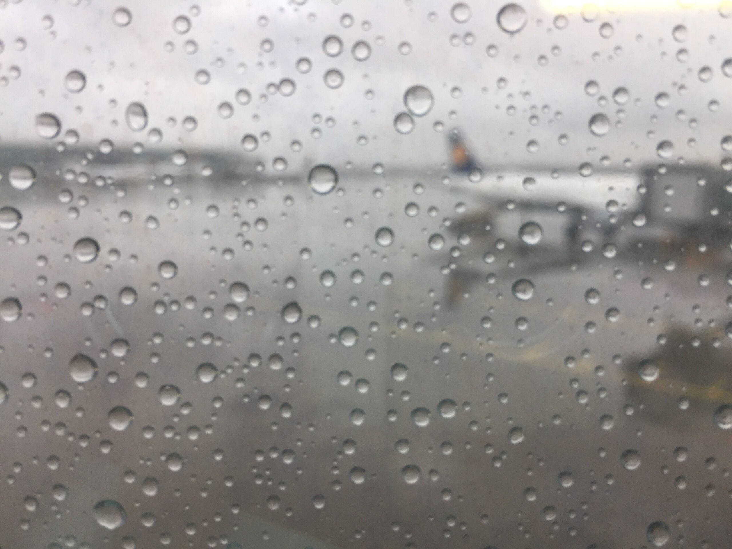 Vue depuis le hublot d'un avion sur une piste d'aéroport et un avion. La mise au point est sur les gouttes d'eau de la vitre. Ce qui est en arrière plan est flou et gris.