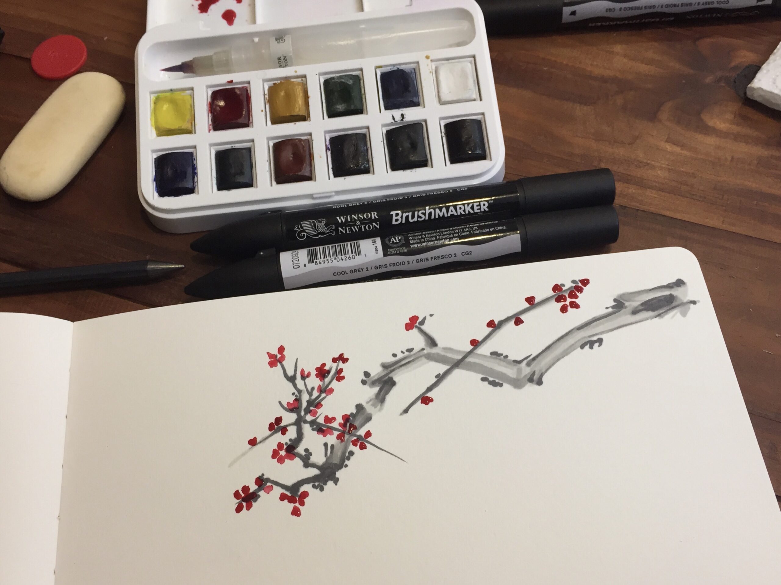 Branch of a blooming tree, done using grey Brushmarkers and alizarin crimson watercolor. The artwork is on a wide drawing book on a table, next to the pens and paint.