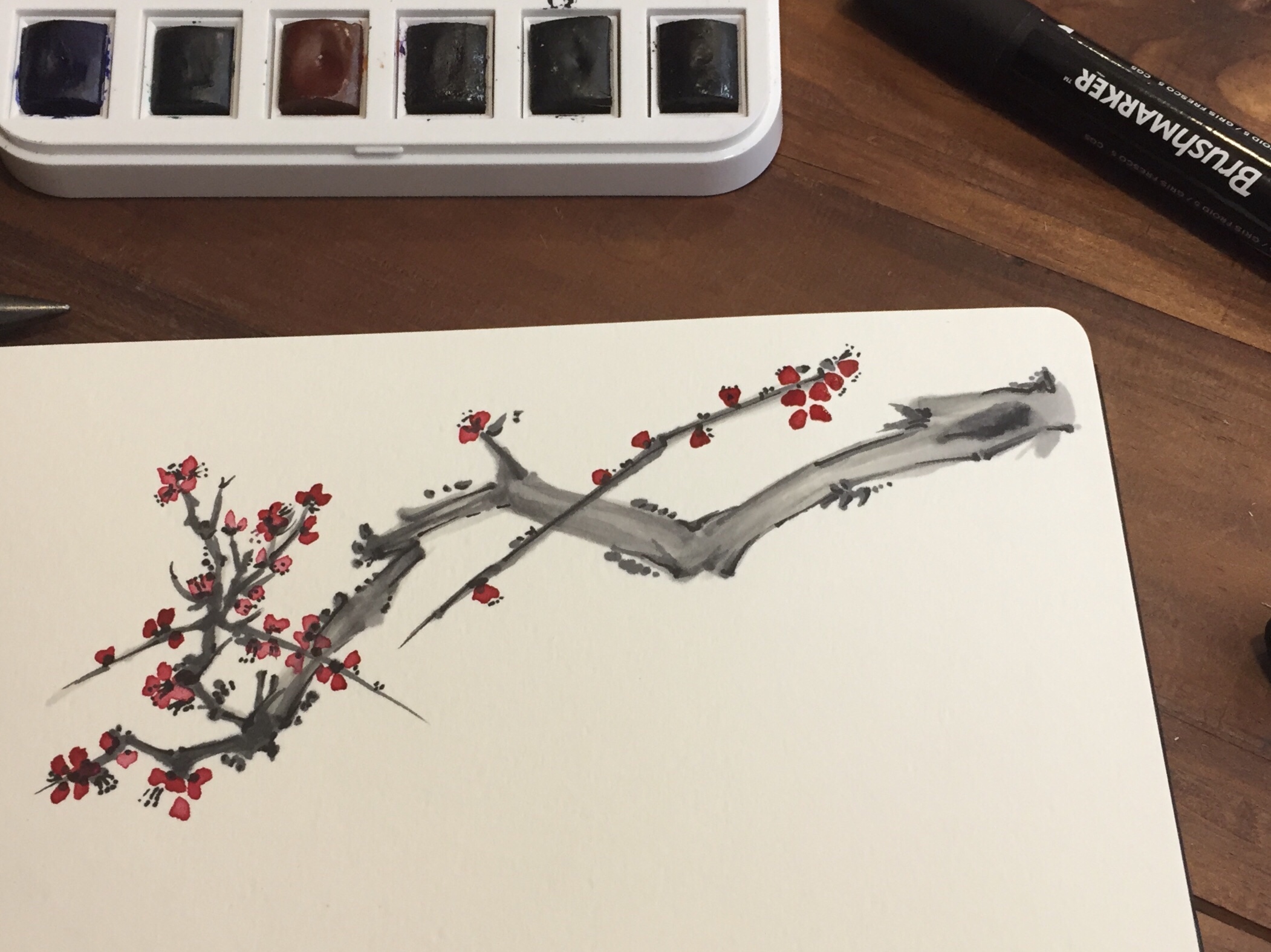 Branch of a blooming tree, to which I added a few black spots. The artwork is on a wide drawing book on a table, next to pens and paint.