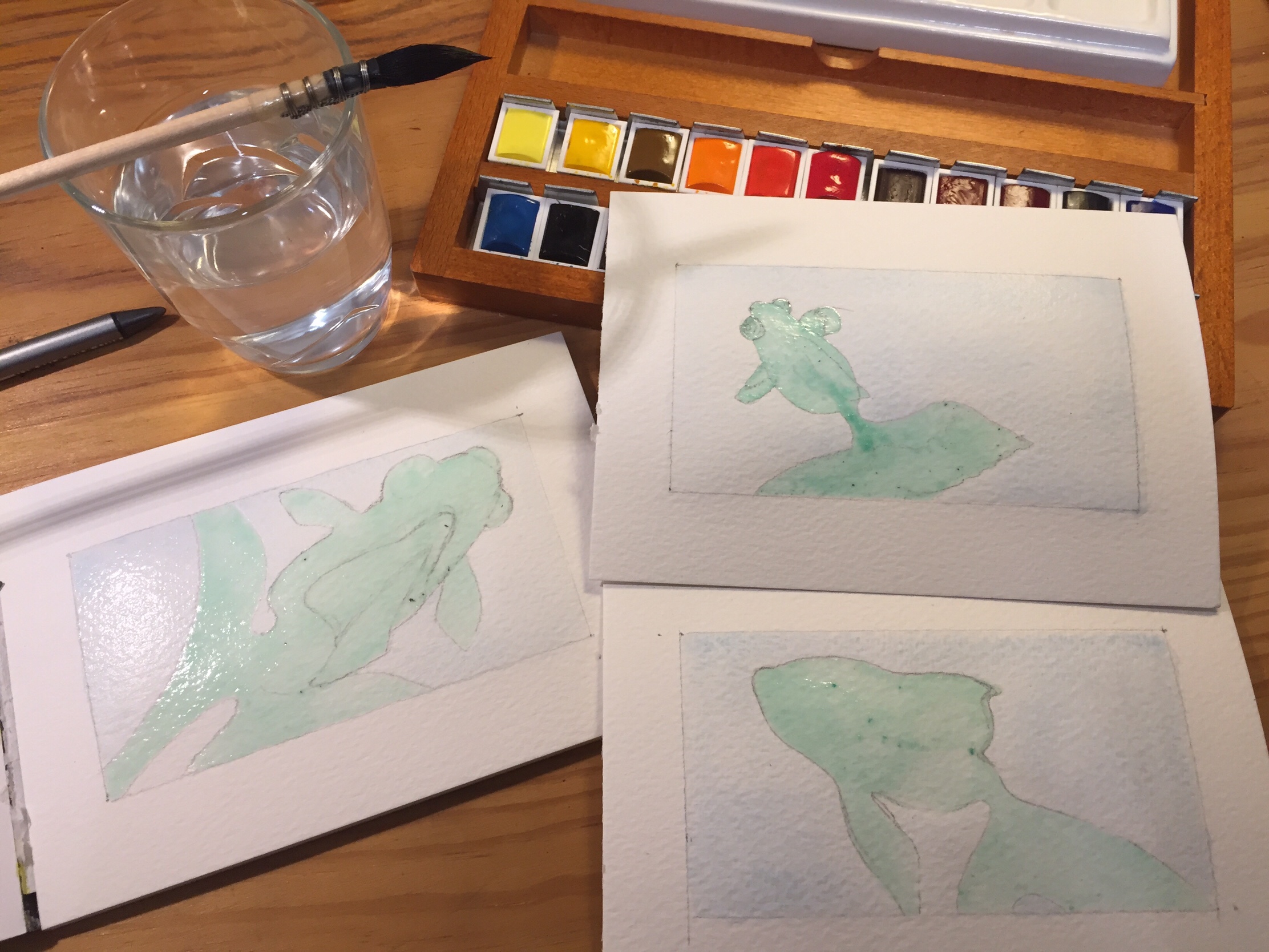 Three postcard sized watercolour sheets where I drew the shapes of goldfish and painted the background in light blue. The watercolour pans, brush and water glass are visible.