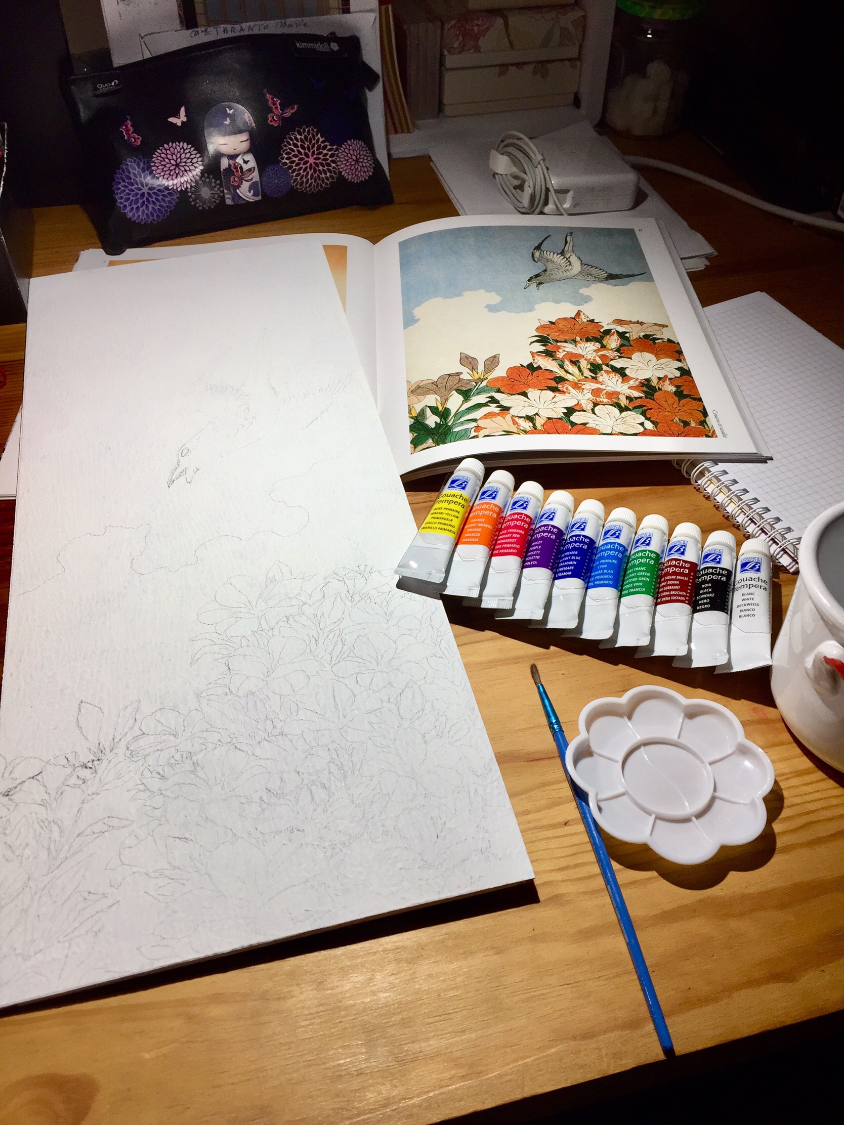 Large white wooden board on a table next to gouache paint tubes, a brush and tiny plastic palette. A book is open at the illustration that I use as reference. A precise pencil sketch is visible on the board.