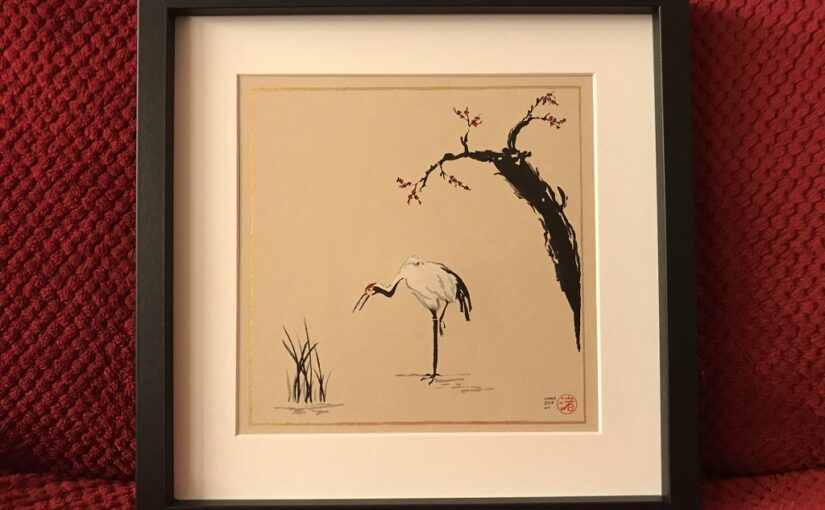 Black square frame and white mount card around a black, grey, white and red ink drawing on taupe toned paper of a Japanese crane standing between some reed and an old tree with red blossoms.
