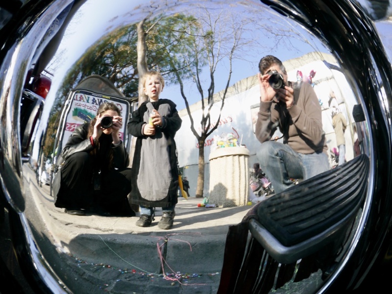 Reflection in a motorcycle part. A small boy stands between a woman (me) and a man crouching and kneeling behind their cameras