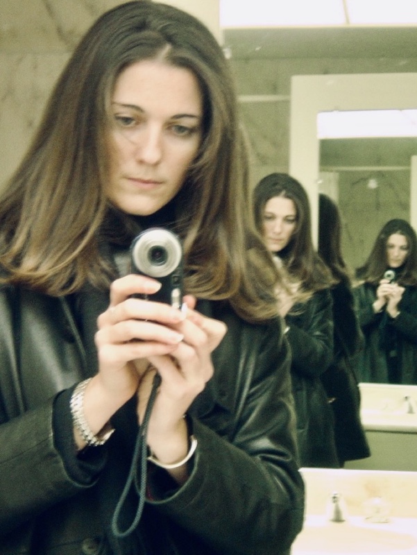 Photo of me, young woman with straight shoulder-length brown hair, reflected in the mirrors of an elevator. I'm holding a dark blue camera in both hands and looking at the screen. I'm wearing a leather coat. I'm reflected three times.