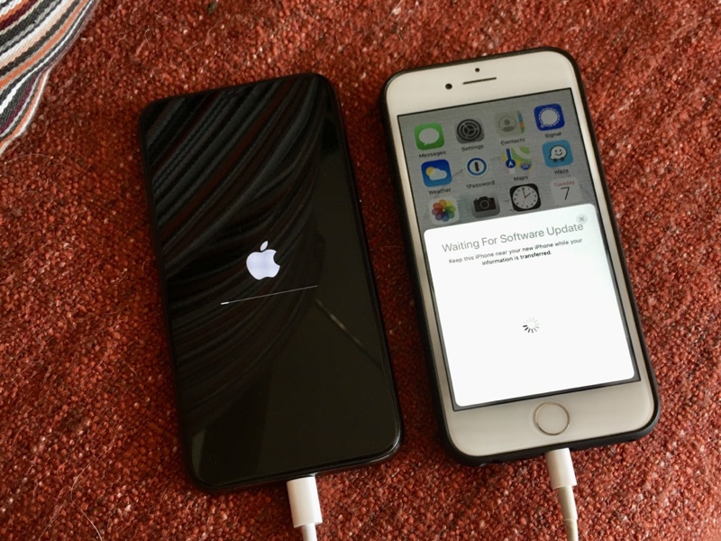 Photo of two iphones side by side, being charged, on a red blanket. One is showing the white Apple logo and is updating, the other is waiting for software update.