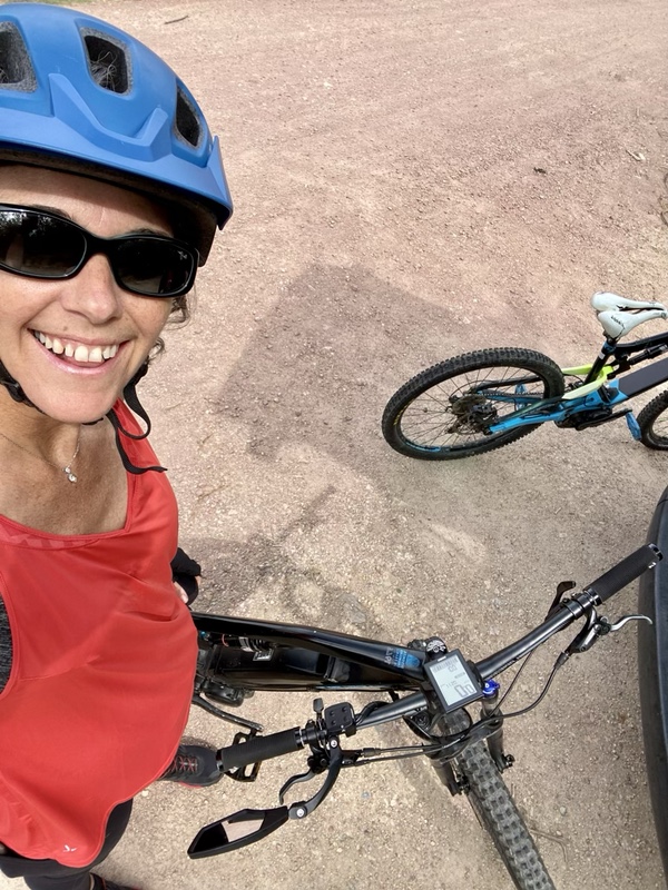 Photo of smiling me by me as I am on a black electric mountain bike, wearing a blue helmet, sunglasses and a red tank top. There is another e-mtb visible next to me.