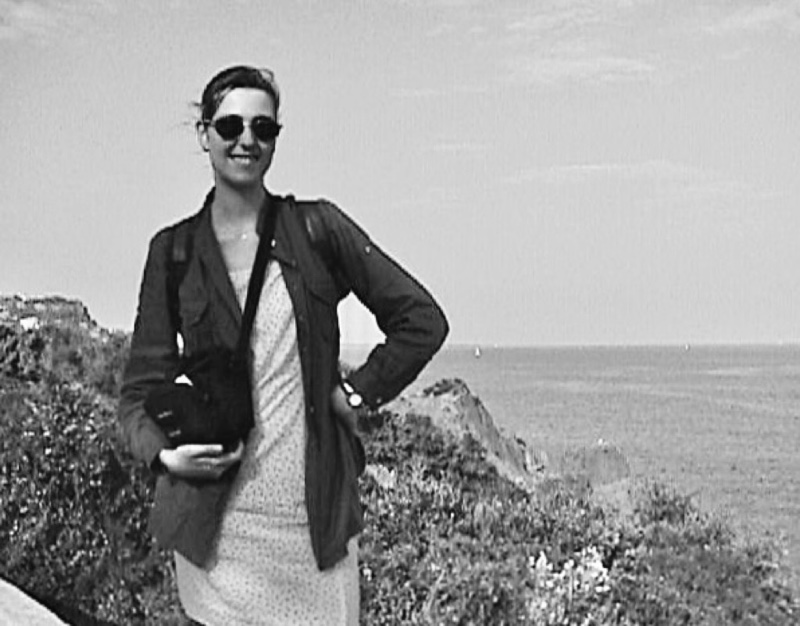 Black and white grainy photo of me, young white woman in sunglasses, holding a camera bag. I stand by vegetation with the sea in my background. My hair is tied up, I wear a flowery light short dress and an open dark shirt.