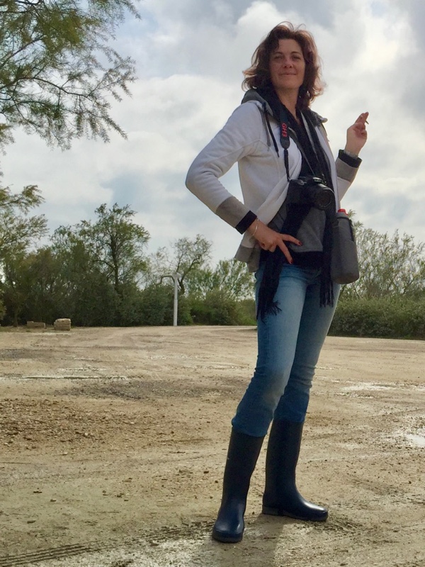Photo of me, smiling woman with short curly hair, standing up in rubber boots and blue jeans on wet sandy ground. I am stylishly smoking while I carry a water bottle and my camera around my neck.