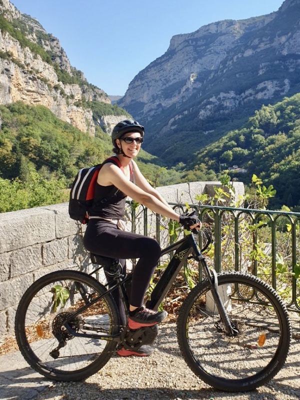 Photo of me smiling as I sit on the electric mountain bike by the end of a former railway bridge. I'm wearing a black tank top and black and red backpack.