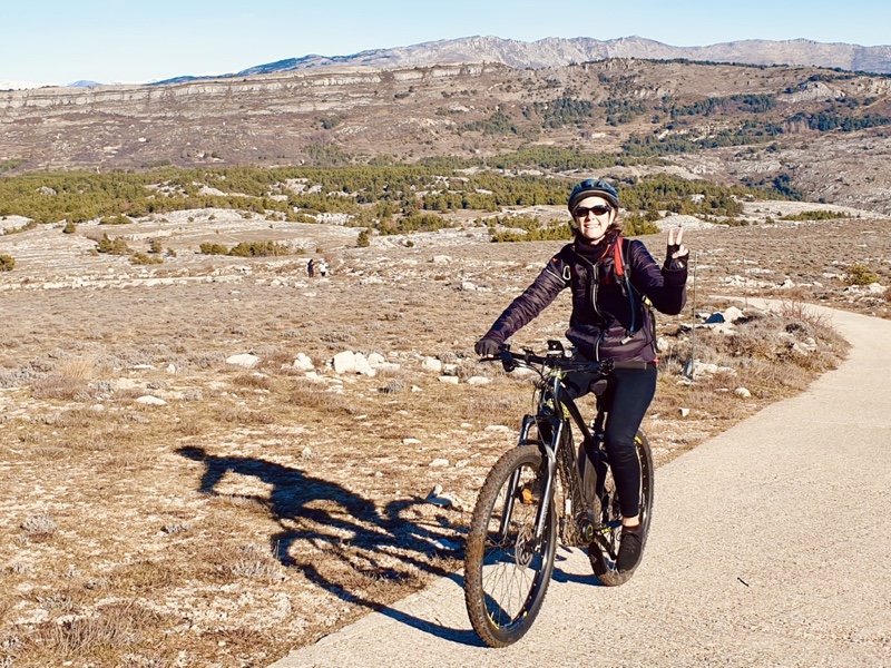 Photo of me riding my electric mountain bike and doing a peace sign on a sunny day. I'm wearing a warm sports coat. The area around is a plateau with rocks and dry grass.