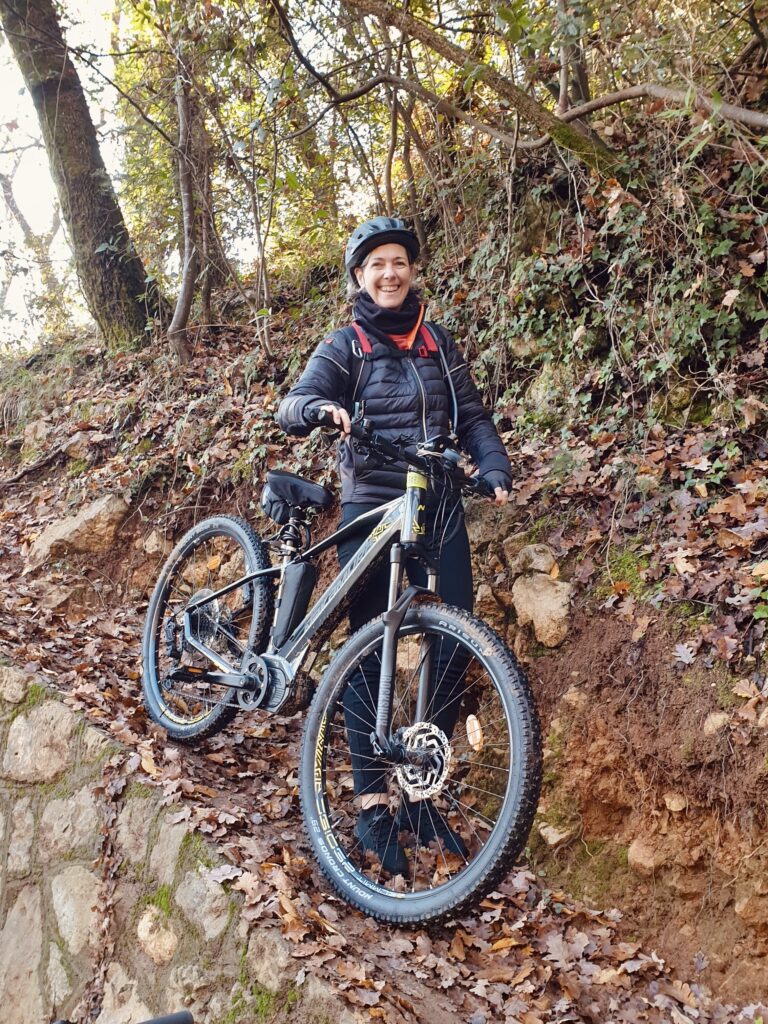 Smiling middle-aged woman wearing a bike helmet standing next to a dark grey mountain bike in a steep incline covered in dead leaves