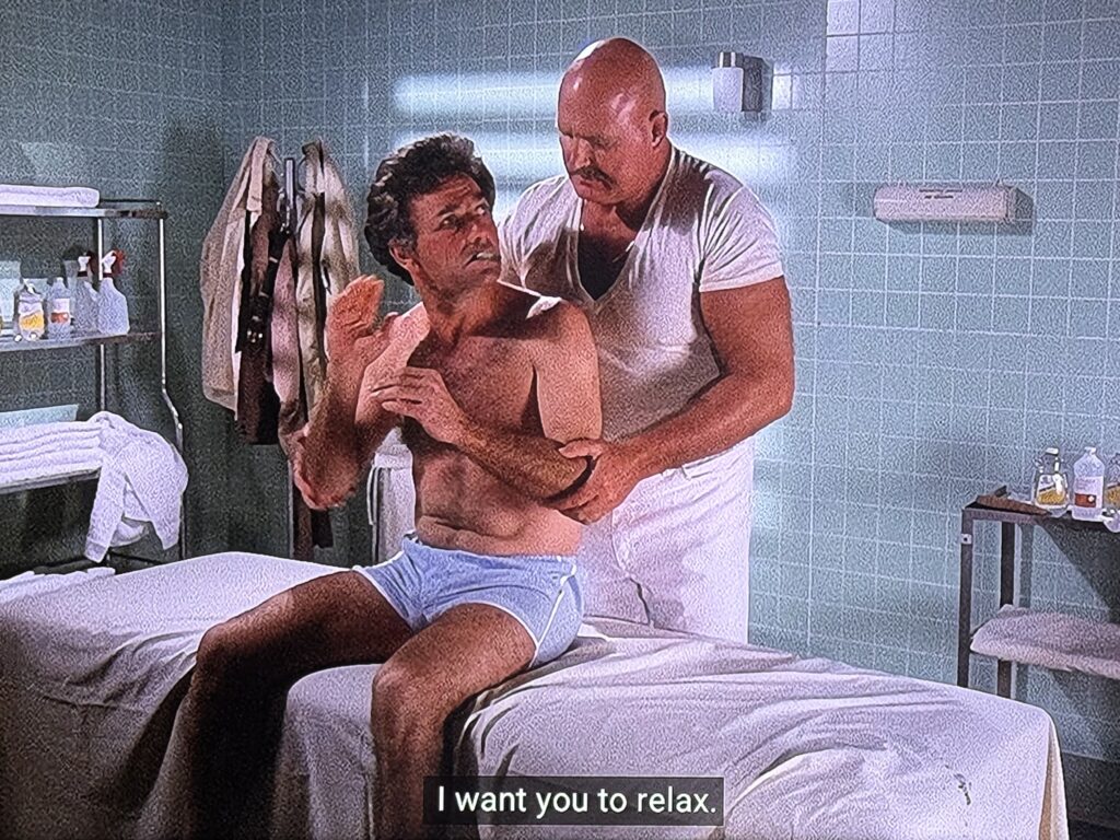 Lieutenant Columbo in boxer shorts, his green cigar at his mouth, seats on a massage table in front of a bald and very muscular man in white telling him “I want you to relax.”
